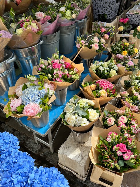 An outdoor display of fresh flower bouquets arranged in metal buckets and wrapped in brown paper, featuring a variety of pastel-colored roses, hydrangeas, and carnations. The bouquets are neatly arran