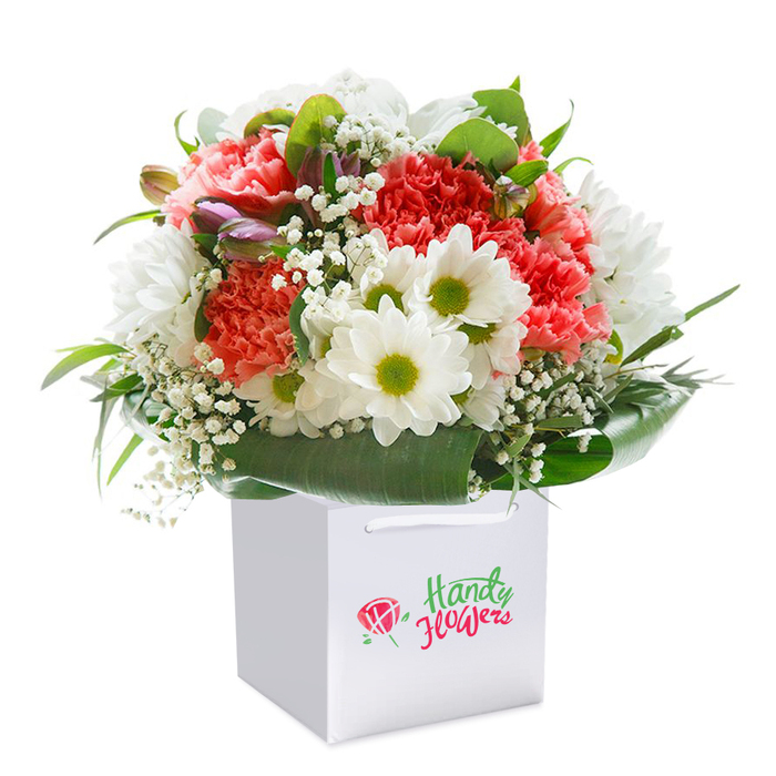 Bursting with the spirit of Whetstone, this striking arrangement unites classic floristry with contemporary elegance. Ruffled coral and red carnations provide a foundation of warm, spirited colour, their textures lush and inviting to the touch. Crisp white daisies, each featuring a gleaming lime-yellow center, nestle harmoniously alongside, imparting a sense of optimism as bright as a sunny morning near Oakleigh Park South. Wisps of baby's breath, soft and ethereal, waft throughout, softening the overall effect, while glimpses of unopened, purple-tipped buds lend a layer of quiet anticipation. Encircling the entire bouquet, abundant deep green foliage fans out in a glossy, natural wrap, giving fullness and stability to the rounded, flourishing shape-a signature of florists who know their Whetstone roots. Sitting gracefully within a spotlessly white, square gift bag, enhanced by sturdy ribbon handles for easy delivery or gifting, the ensemble is positioned against a crisp white background that highlights every colour and contour. Bright, diffused light brings out the freshness and vibrancy of each flower, emphasizing this bouquet's readiness for any celebration across Whetstone. Whether for birthdays, anniversaries, or simply a spontaneous gesture of kindness, this arrangement brings the thoughtful artistry of Whetstone's floral community directly to the hands and homes of local residents.