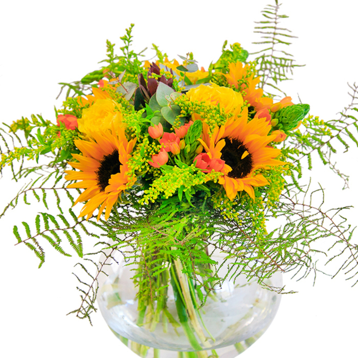 As if bathed in the golden afternoon light of a Whetstone garden, this bouquet is a heartening vision of warmth and elegance. Towering sunflowers with inky brown centers are arranged to face outward, spreading their vibrant yellow petals like rays across a landscape of blooms. Nestled alongside are clusters of densely petaled yellow roses, each unfurling with classic grace, their plush surfaces whispering of timeless romance. Sprigs of small, fiery red Hypericum berries punctuate the design, their colour dancing harmoniously among a bed of fresh green ferns and delicate filler sprigs. Feathery foliage spills over the rounded edges of the bouquet, softening and balancing the boldness of the sunflowers with a sense of natural movement. Eucalyptus leaves-cool and silvery-peek from within, their scent reminiscent of peaceful strolls through Whetstone's leafy lanes. The arrangement is housed in a clear glass vase, water brimming and stems visible, offering an impression of pure freshness and modern artistry. Light plays gently over the flowers, enhancing their textures and hues. This bouquet, whether chosen to mark a milestone or to simply enliven a kitchen table in Whetstone, is both a visual feast and a celebration of nature's abundance, crafted with expert care and available for local delivery.